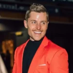 A professional portrait of Sawyer Hemsley smiling in a vibrant orange blazer at a formal event.
