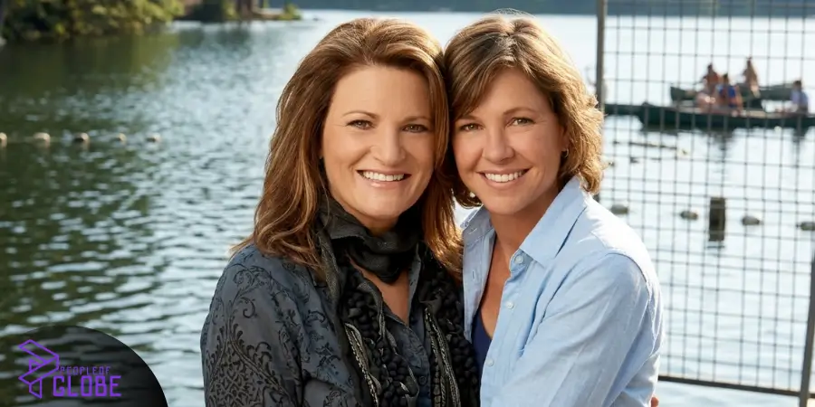 A portrait photo of Martie Allen and her partner Kristy McNichol standing together smiling, with a scenic lake and outdoor setting in the background.